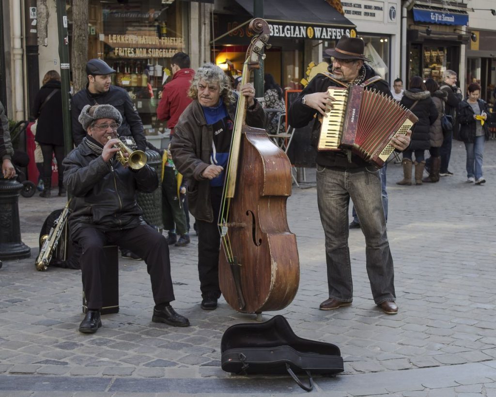 Sokakta akordeon ve keman çalan iki müzisyen, etraflarında pastel tonlu eski binalar ve birkaç izleyici